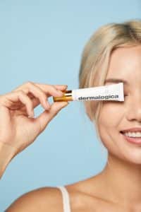 What causes dark circles under the eyes can make us to want to hide them. In this image a woman is holding an eye cream up to her eye.
